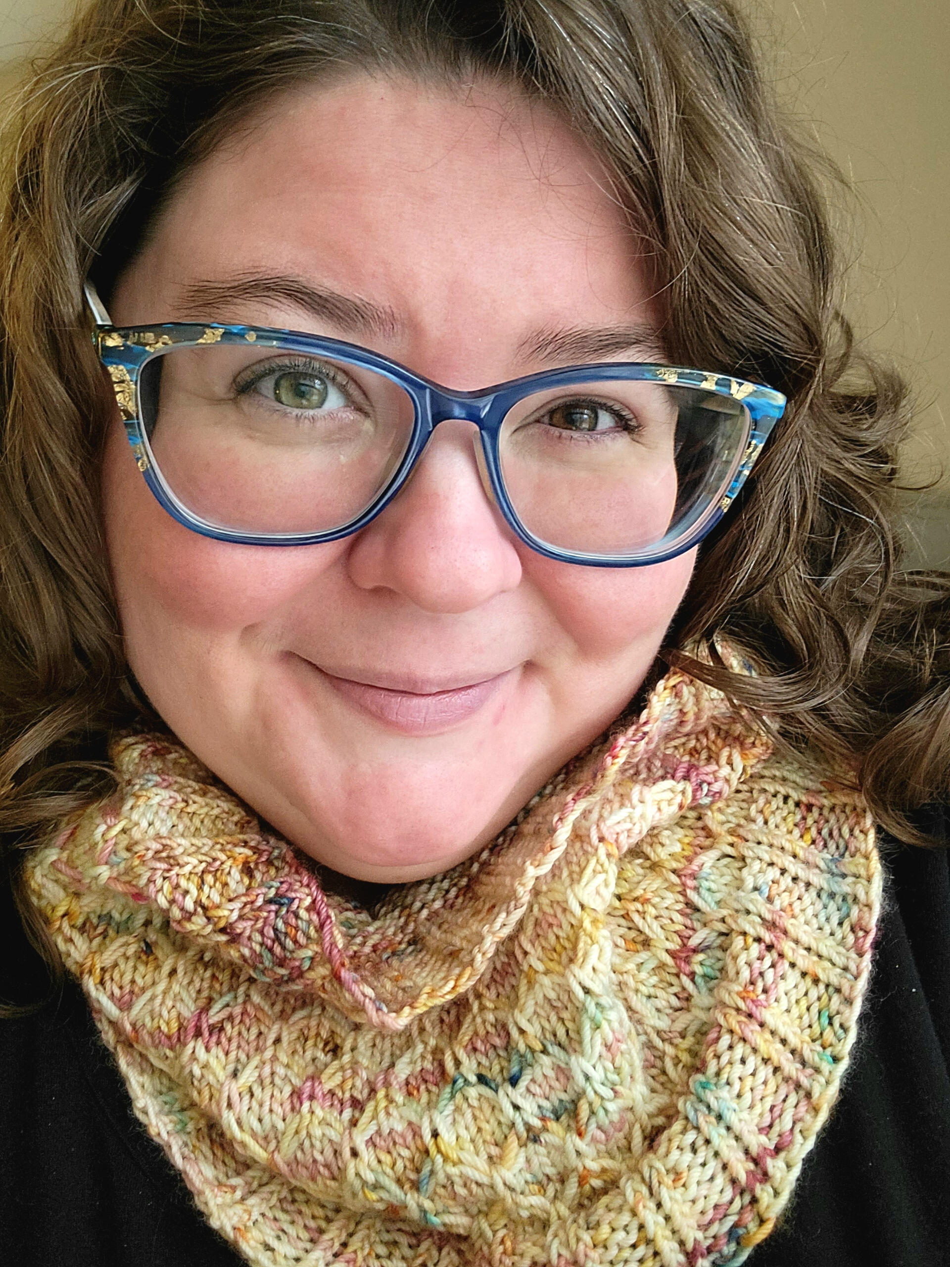 A smiling white woman with oversized blue glasses, curling brown hair, and hazel eyes. She wears a knitted cowl with various shades of gold and speckled with autumnal colors.