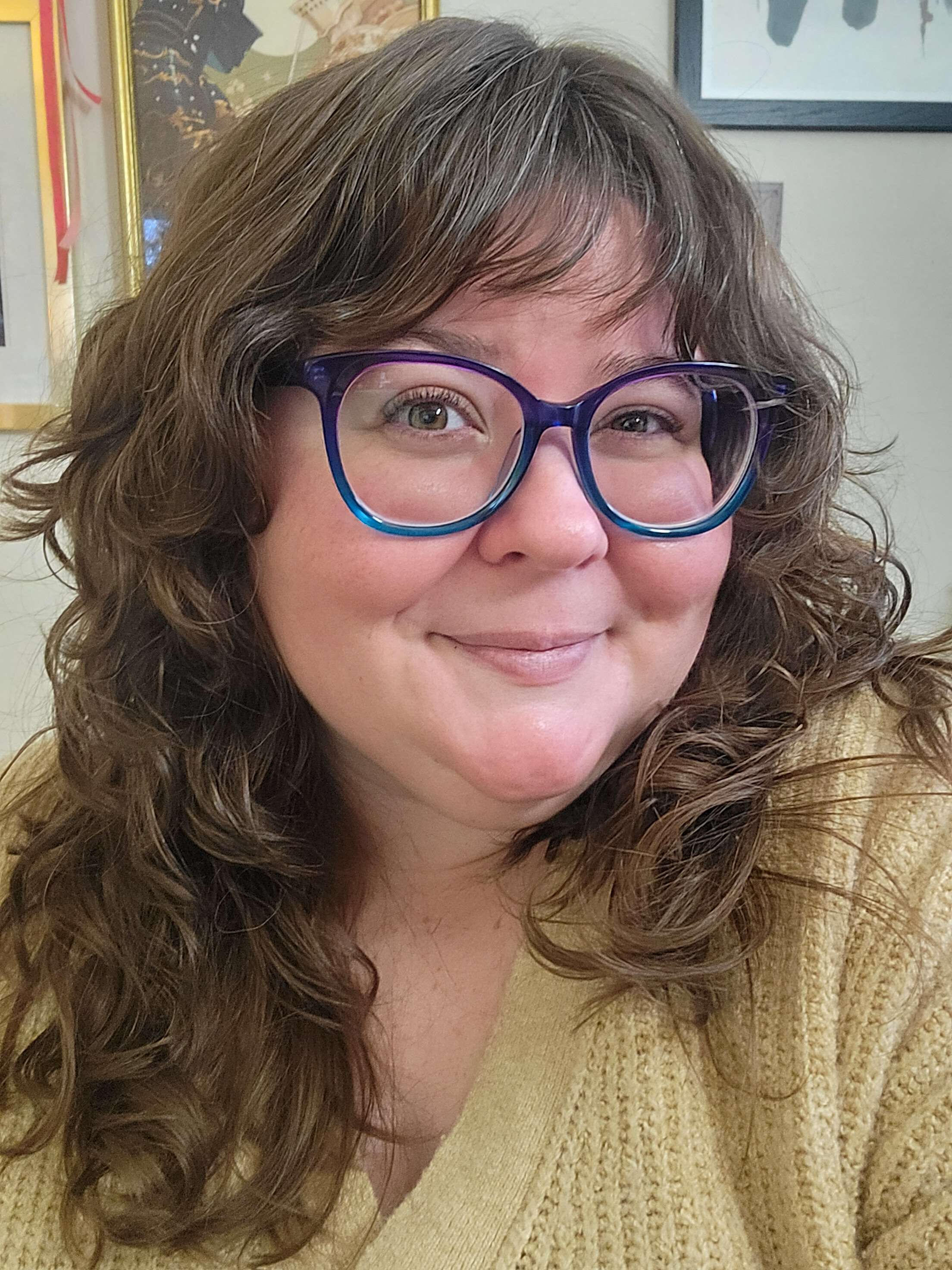 A smiling white woman with oversized blue glasses, curling brown hair, and hazel eyes. She wears a gold sweater, and is seated in front of a wall featuring several pieces of framed art.